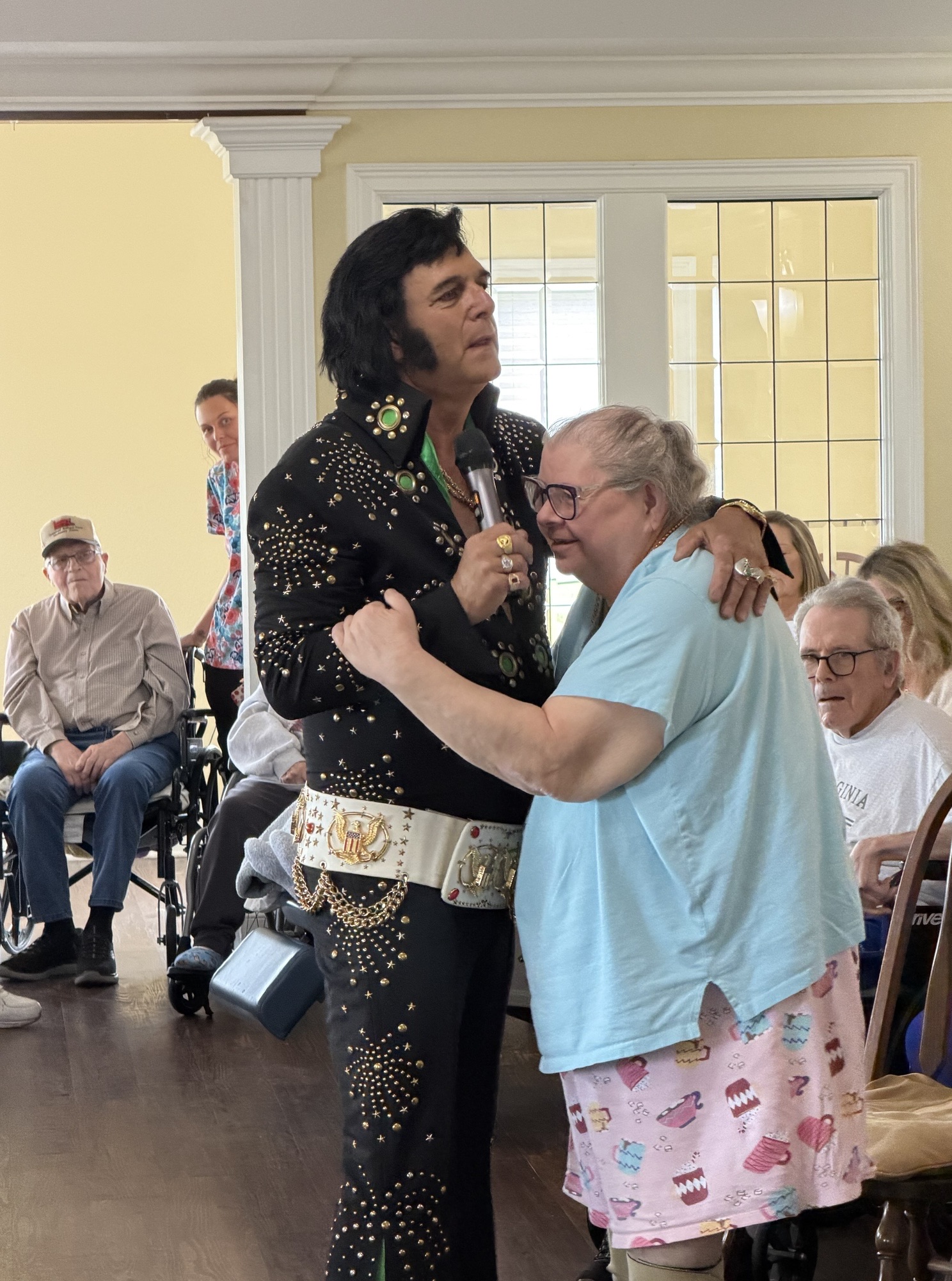 Elvis tribute artist in a jeweled black jumpsuit sings into a microphone as a woman dances beside him; seated audience watches in a bright room.