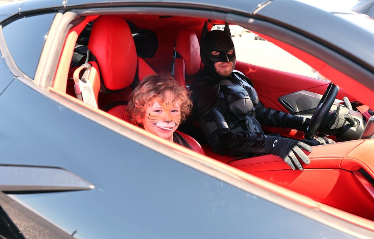 Child with tiger face paint smiling in a red sports car, seated beside a Batman-costumed driver in the driver's seat.