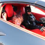Child with tiger face paint smiling in a red sports car, seated beside a Batman-costumed driver in the driver's seat.