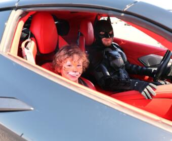 Child with tiger face paint smiling in a red sports car, seated beside a Batman-costumed driver in the driver's seat.