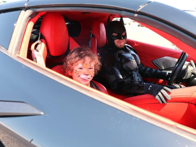 Child with tiger face paint smiling in a red sports car, seated beside a Batman-costumed driver in the driver's seat.