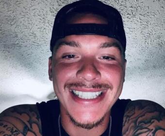 Close-up of a smiling man wearing a backward black cap, with visible arm tattoos in a sleeveless shirt.