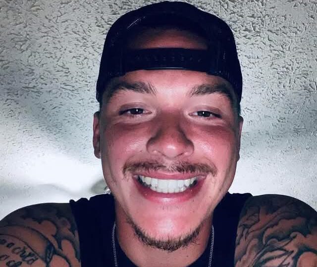 Close-up of a smiling man wearing a backward black cap, with visible arm tattoos in a sleeveless shirt.