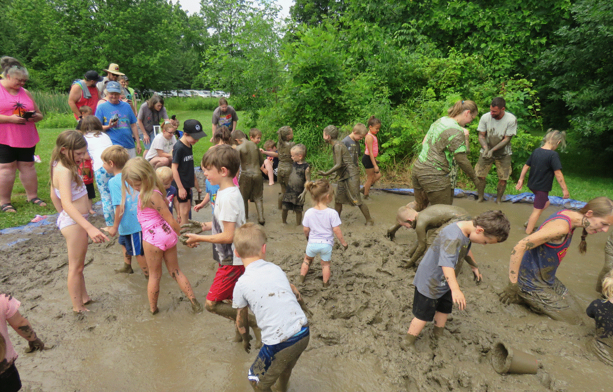 Group of children and adults playing in deep mud outdoors, throwing mud and wading in a large muddy pit surrounded by green trees.