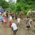 Group of children and adults playing in deep mud outdoors, throwing mud and wading in a large muddy pit surrounded by green trees.