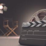 Clapperboard in the foreground with a film reel, set on a studio backdrop with a bright spotlight and blurred director chairs in the background.