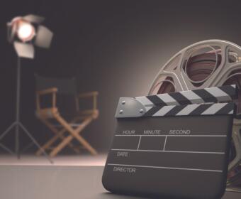 Clapperboard in the foreground with a film reel, set on a studio backdrop with a bright spotlight and blurred director chairs in the background.