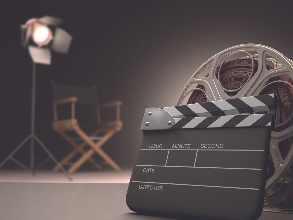 Clapperboard in the foreground with a film reel, set on a studio backdrop with a bright spotlight and blurred director chairs in the background.