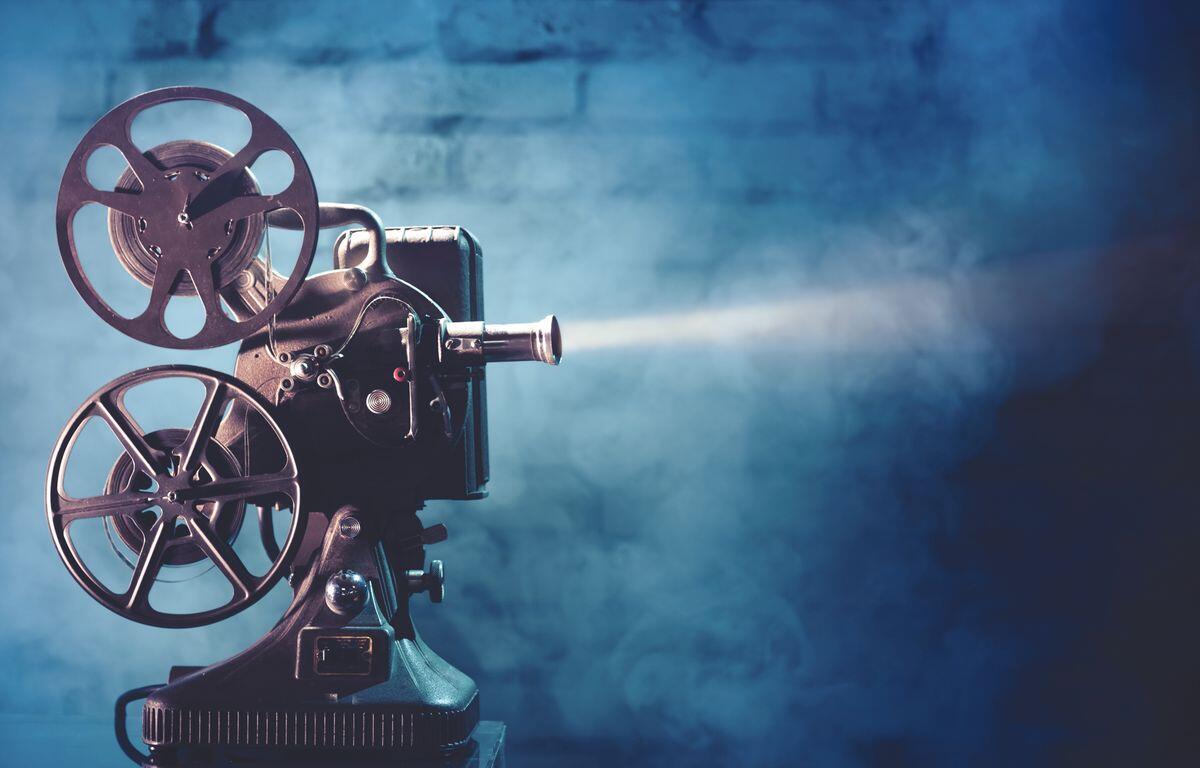 Vintage film projector with two reels and lens, casting a beam of light against a blue studio backdrop.