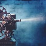 Vintage film projector with two reels and lens, casting a beam of light against a blue studio backdrop.