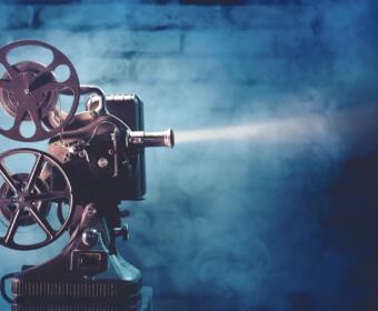 Vintage film projector with two reels and lens, casting a beam of light against a blue studio backdrop.