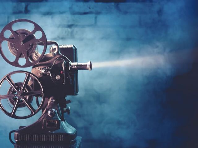Vintage film projector with two reels and lens, casting a beam of light against a blue studio backdrop.
