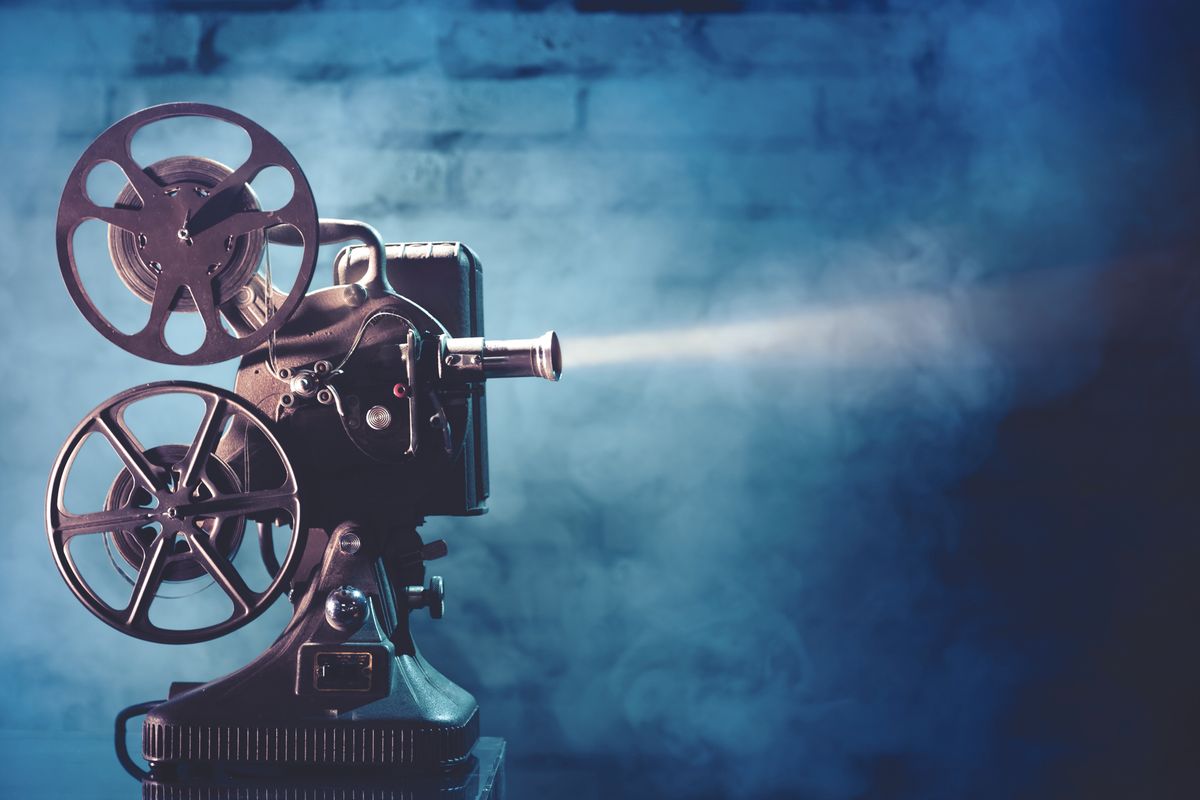 Vintage film projector with two reels and lens, casting a beam of light against a blue studio backdrop.