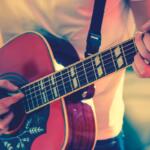 Close-up of a person’s hands playing an acoustic guitar, fingers on the fretboard and strumming near the soundhole.