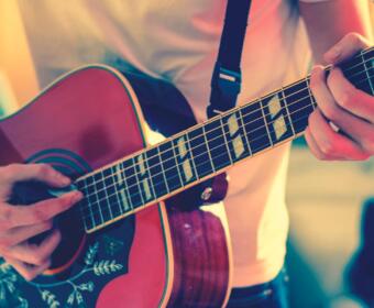 Close-up of a person’s hands playing an acoustic guitar, fingers on the fretboard and strumming near the soundhole.