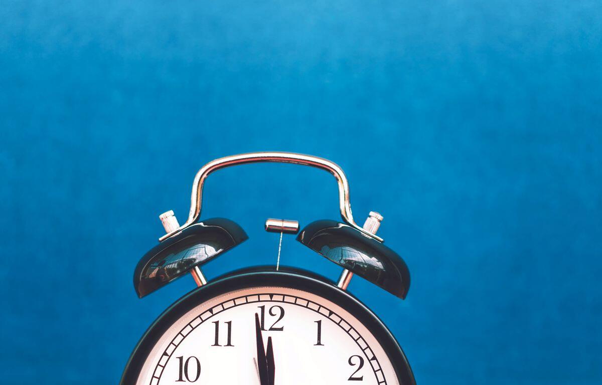 Analog alarm clock with twin bells against a solid blue background, showing 12:00.