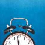Analog alarm clock with twin bells against a solid blue background, showing 12:00.