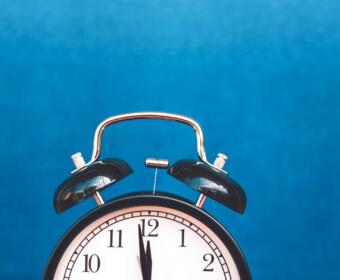 Analog alarm clock with twin bells against a solid blue background, showing 12:00.