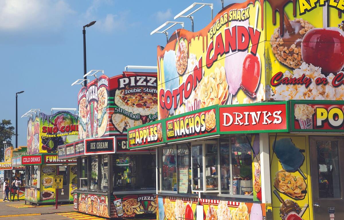 Row of bright carnival food stands with large signs for cotton candy, nachos, pizza, ice cream and drinks.