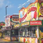 Row of bright carnival food stands with large signs for cotton candy, nachos, pizza, ice cream and drinks.