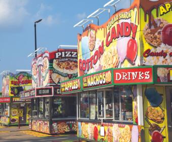 Row of bright carnival food stands with large signs for cotton candy, nachos, pizza, ice cream and drinks.