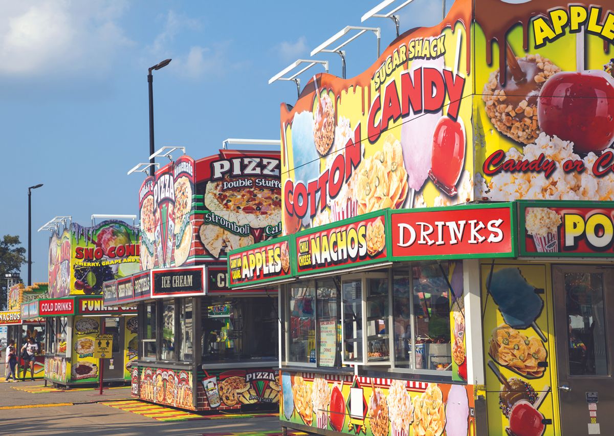 Row of bright carnival food stands with large signs for cotton candy, nachos, pizza, ice cream and drinks.