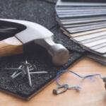 Hammer and nails on a textured mat with color swatches, blue string, and a small metal heart charm on a wooden work surface.