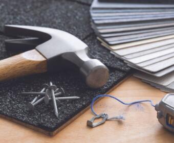 Hammer and nails on a textured mat with color swatches, blue string, and a small metal heart charm on a wooden work surface.