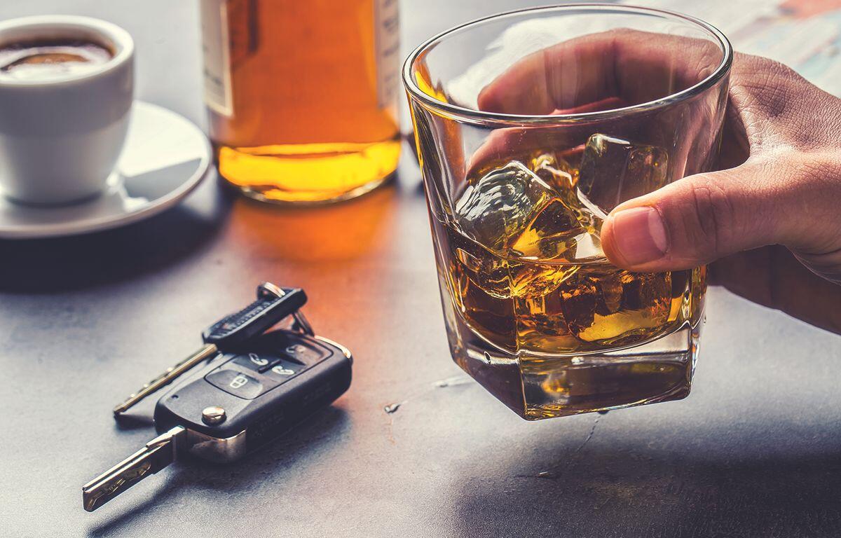 Hand holding a glass of whiskey with ice on a table; a bottle, car keys, and a cup of coffee visible in the background (implies drinking and driving risk).