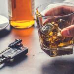 Hand holding a glass of whiskey with ice on a table; a bottle, car keys, and a cup of coffee visible in the background (implies drinking and driving risk).