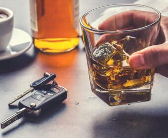 Hand holding a glass of whiskey with ice on a table; a bottle, car keys, and a cup of coffee visible in the background (implies drinking and driving risk).