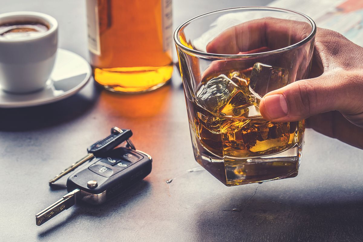Hand holding a glass of whiskey with ice on a table; a bottle, car keys, and a cup of coffee visible in the background (implies drinking and driving risk).
