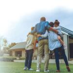 Family of four hugs in front of a house with a for-sale sign on the lawn, celebrating a purchase or sale