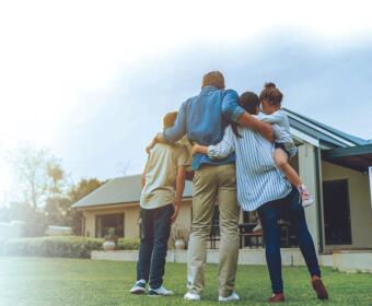 Family of four hugs in front of a house with a for-sale sign on the lawn, celebrating a purchase or sale