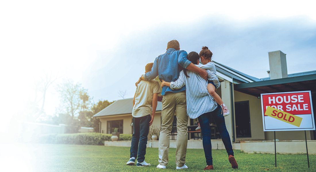 Family of four hugs in front of a house with a for-sale sign on the lawn, celebrating a purchase or sale