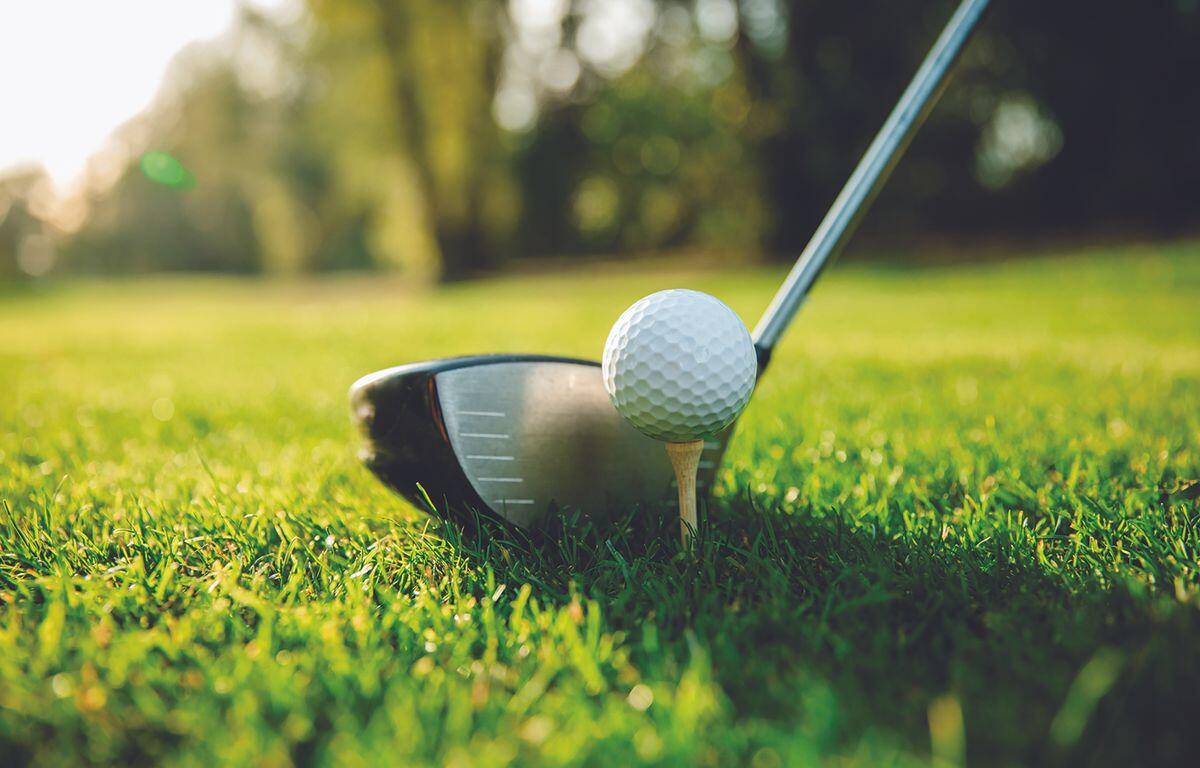 Golf ball teed up on a tee with a driver behind it on a sunny, grassy golf course.