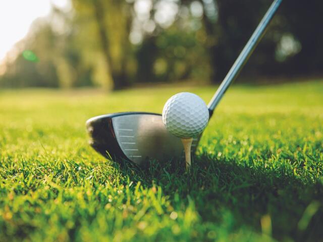 Golf ball teed up on a tee with a driver behind it on a sunny, grassy golf course.