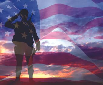 Silhouette of a soldier saluting with a rifle, overlaid on a waving American flag at sunset/dusk Вост.