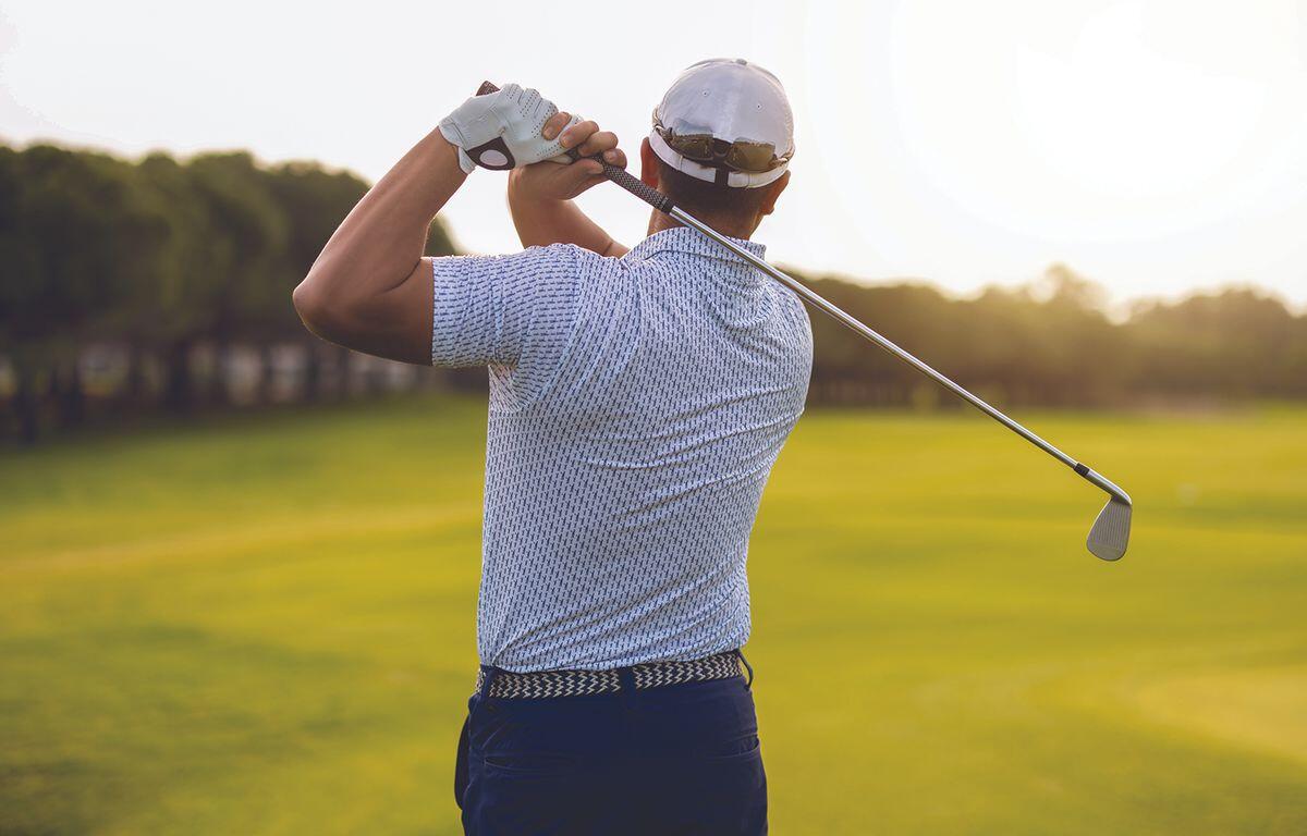 Golfer mid backswing on a sunny golf course, seen from behind with a white cap and gloved hand.
