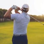 Golfer mid backswing on a sunny golf course, seen from behind with a white cap and gloved hand.