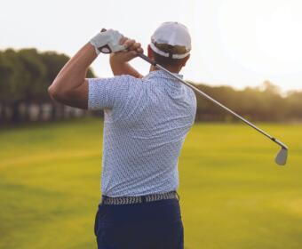 Golfer mid backswing on a sunny golf course, seen from behind with a white cap and gloved hand.