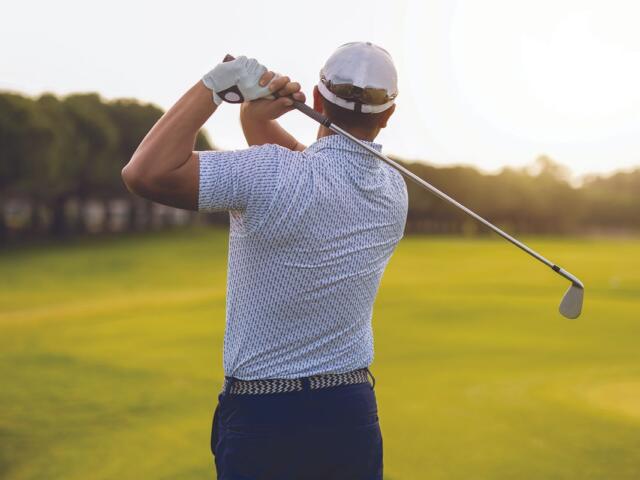 Golfer mid backswing on a sunny golf course, seen from behind with a white cap and gloved hand.