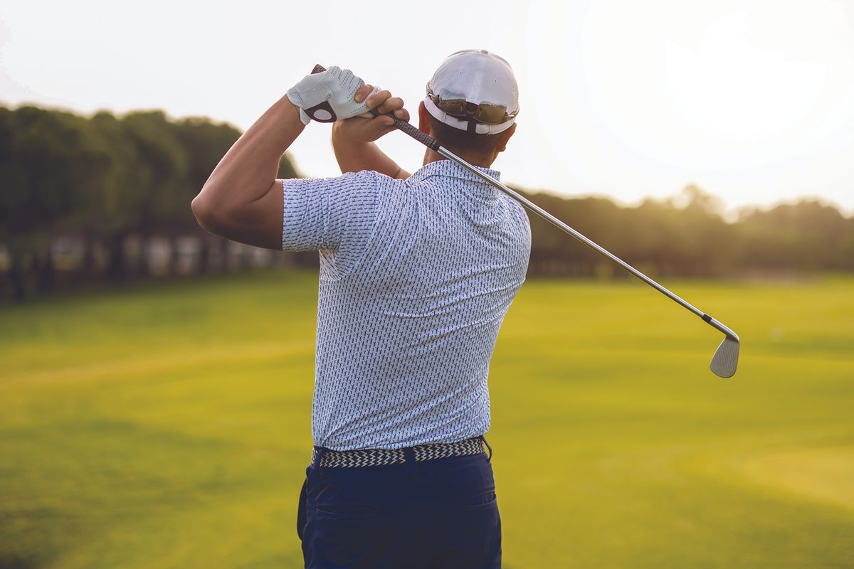 Golfer mid backswing on a sunny golf course, seen from behind with a white cap and gloved hand.
