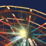 Blurred ferris wheel at night with multicolored lights radiating from the center around a bright glow at the hub.