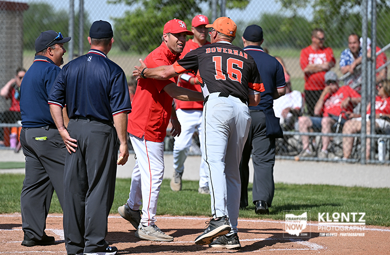 Baseball players argue at home plate as umpires converse, with spectators behind a chain-link fence.