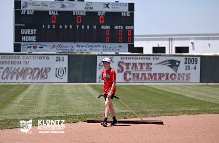 Young person in a red shirt and helmet walks on the infield with a drag mat on a baseball/softball field; scoreboard and 'State Champions' banners visible in background.
