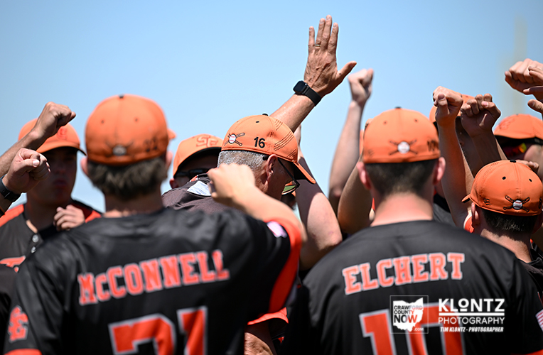 Group of football players in orange caps and black jerseys celebrate in a huddle, raising their fists and hands in the air; names McDonnell and Elchert appear on the backs.