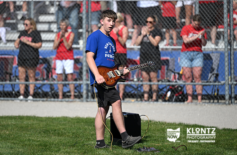 Teenage boy in a blue shirt playing an electric guitar on a grass field at a fenced sports event, with spectators behind him.