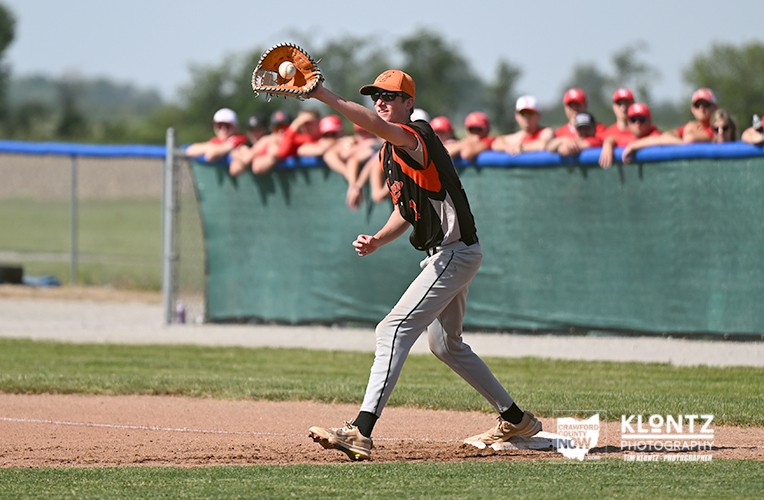 Baseball pitcher in an orange cap throws a pitch on the mound during a game, with teammates watching behind the fence.