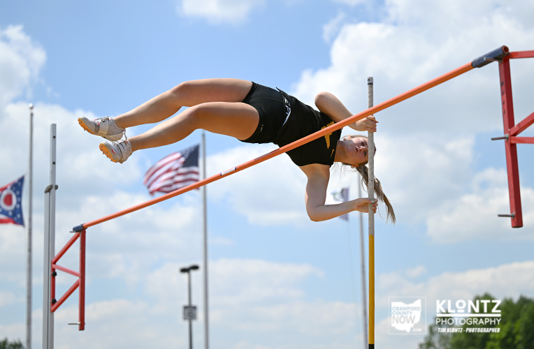 Female pole vaulter in a black outfit clears an orange bar mid-air outdoors against a blue sky with flags in the background.